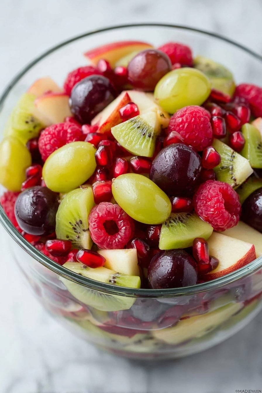A clear glass bowl filled with a colorful mix of fresh fruits on a white marbled surface. The fruit salad has three main layers: the bottom layer is mixed with light green apple pieces with a smooth texture and thin red skin, the middle layer features bright green and dark purple grapes that are oval and shiny, and the top layer is scattered with vibrant red raspberries that have a soft, bumpy texture and small pomegranate seeds adding shiny ruby red dots. The bowl is shown from above, with a lime slice partially visible at the top edge of the image and some loose raspberries and grapes scattered around the bowl. Photo taken with an iphone --ar 2:3 --v 7 - Festive Christmas Fruit Salad, holiday fruit salad, Christmas fruit salad, easy holiday desserts, healthy Christmas treat