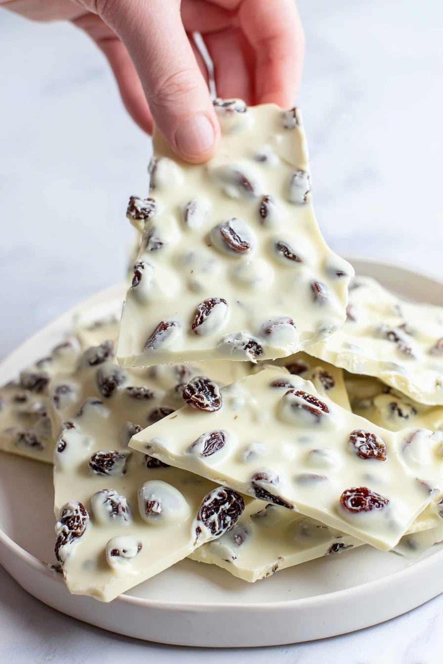The image shows a stack of white chocolate pieces with nuts and dried red cranberries inside. There are about five layers of irregular-shaped white chocolate bars, each studded with golden brown nuts and dark red cranberries. The bars are stacked unevenly on top of each other on a white marbled surface. Some individual nuts and cranberries are scattered loosely around the base of the stack. The background is soft and mostly white, making the white chocolate and colorful nuts and berries stand out clearly. Photo taken with an iphone --ar 2:3 --v 7 - Cranberry Pistachio White Chocolate Bark, holiday white chocolate bark, easy festive treats, quick dessert recipes, crunchy white chocolate snacks
