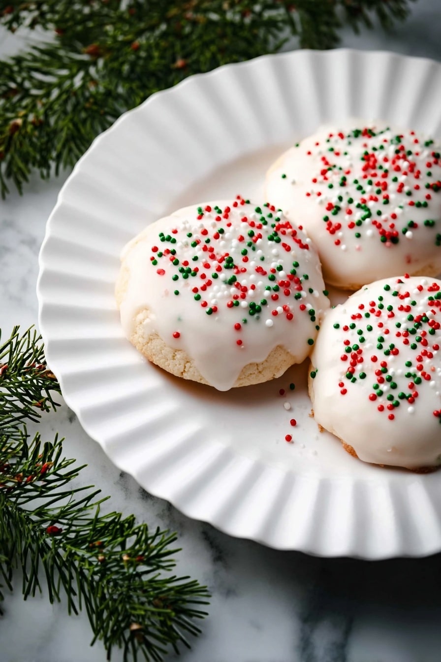 Three round cookies are placed on a large white fluted plate. Each cookie has a thick, smooth white glaze coating on top and is sprinkled with small red, green, and white round sprinkles scattered lightly over the surface. The plate sits on a white marbled surface, with green pine-like branches to the left adding a festive touch. The lighting highlights the shiny glaze texture on the cookies. photo taken with an iphone --ar 2:3 --v 7 - Italian Ricotta Cookies with Glaze, Ricotta cookie recipe, Italian dessert cookies, Cakey ricotta cookies, Easy Italian cookies with glaze