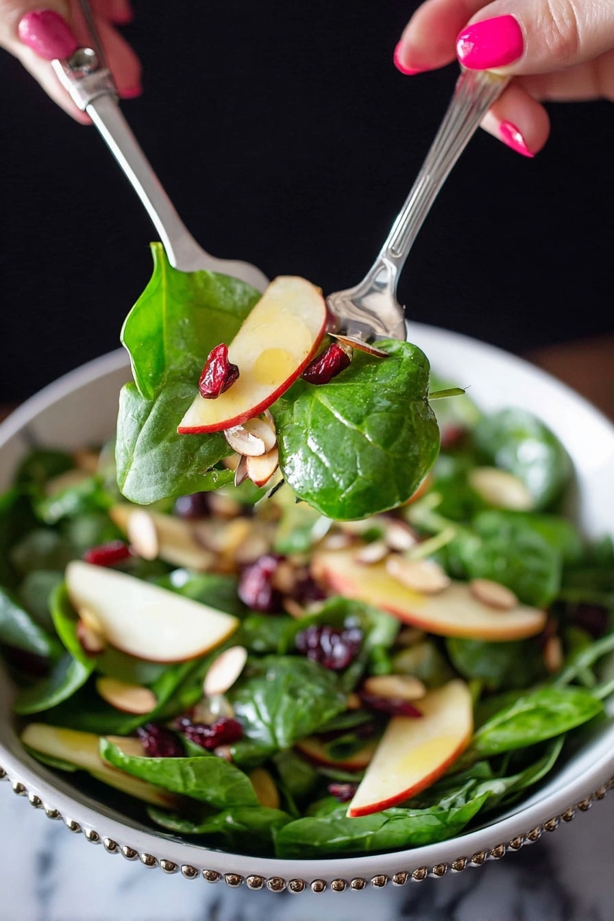 A close-up of a fresh salad being lifted by two silver utensils held by a woman's hand with pink nail polish. The salad has bright green spinach leaves, thin light yellow apple slices with red edges, small dark red dried cranberries, and light brown sliced almonds mixed inside a white bowl with a beaded rim. The background is dark, contrasting with the vibrant colors of the salad, and the surface underneath the bowl is a white marbled texture. photo taken with an iphone --ar 2:3 --v 7 - Apple Cranberry Spinach Salad, healthy fruit and vegetable salad, easy holiday salad recipe, fresh winter salad, nutritious spinach salad