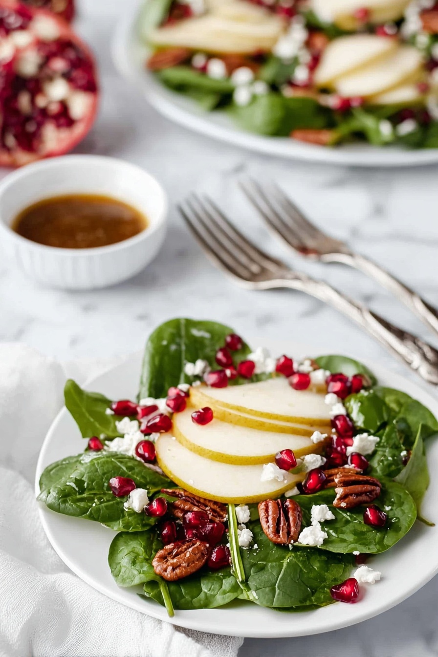 The dish shows a small white plate with a fresh salad made of three main layers. The first bottom layer consists of fresh dark green spinach leaves spread across the plate. On top of this, the second layer has thin round slices of light yellow pear arranged evenly. The third layer is made up of small bright red pomegranate seeds scattered over the pears and spinach, along with small white crumbly pieces of cheese, and a few medium brown pecan nuts. In the background, there is a larger white plate with the same salad and a small white bowl filled with a brown dressing in the center, all set on a white marbled surface. Near the small plate are two silver forks resting on a white cloth. photo taken with an iphone --ar 2:3 --v 7 - Christmas Wreath Salad with Pomegranate and Pear, festive holiday salad, easy Christmas salad recipe, fruit and feta holiday salad, wreath-shaped salad