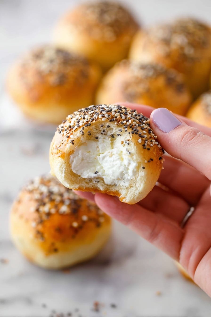 A close-up view of a woman's hand holding a small round bread roll with a golden brown crust topped with a mix of black and white seeds. The roll has a bite taken out, revealing a soft white inside filled with creamy white cheese. In the background, several similar bread rolls are placed on a white marbled surface, each with the same seed topping, slightly out of focus. Photo taken with an iphone --ar 2:3 --v 7 - Stuffed Bagel Bites with Cream Cheese, bagel bite recipes, quick cheese snack, no yeast bagel bites, easy appetizer ideas