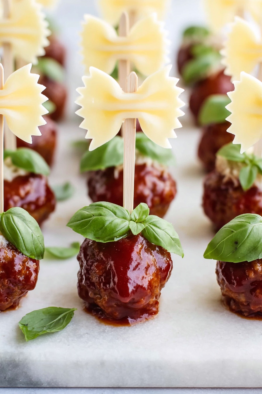 A close-up of a three-layered appetizer on a small wooden stick held by a woman's hand; the bottom layer is a shiny, dark red meatball coated with sauce, the middle layer is a bright green basil leaf with visible veins, and the top layer is a pale yellow bowtie pasta piece with a slightly rough texture and curved edges. The background shows a soft focus white marbled texture. Photo taken with an iphone --ar 2:3 --v 7 - Cocktail Meatball and Bowtie Pasta Skewers, party appetizer ideas, easy meatball skewers, finger food for gatherings, fun pasta skewers