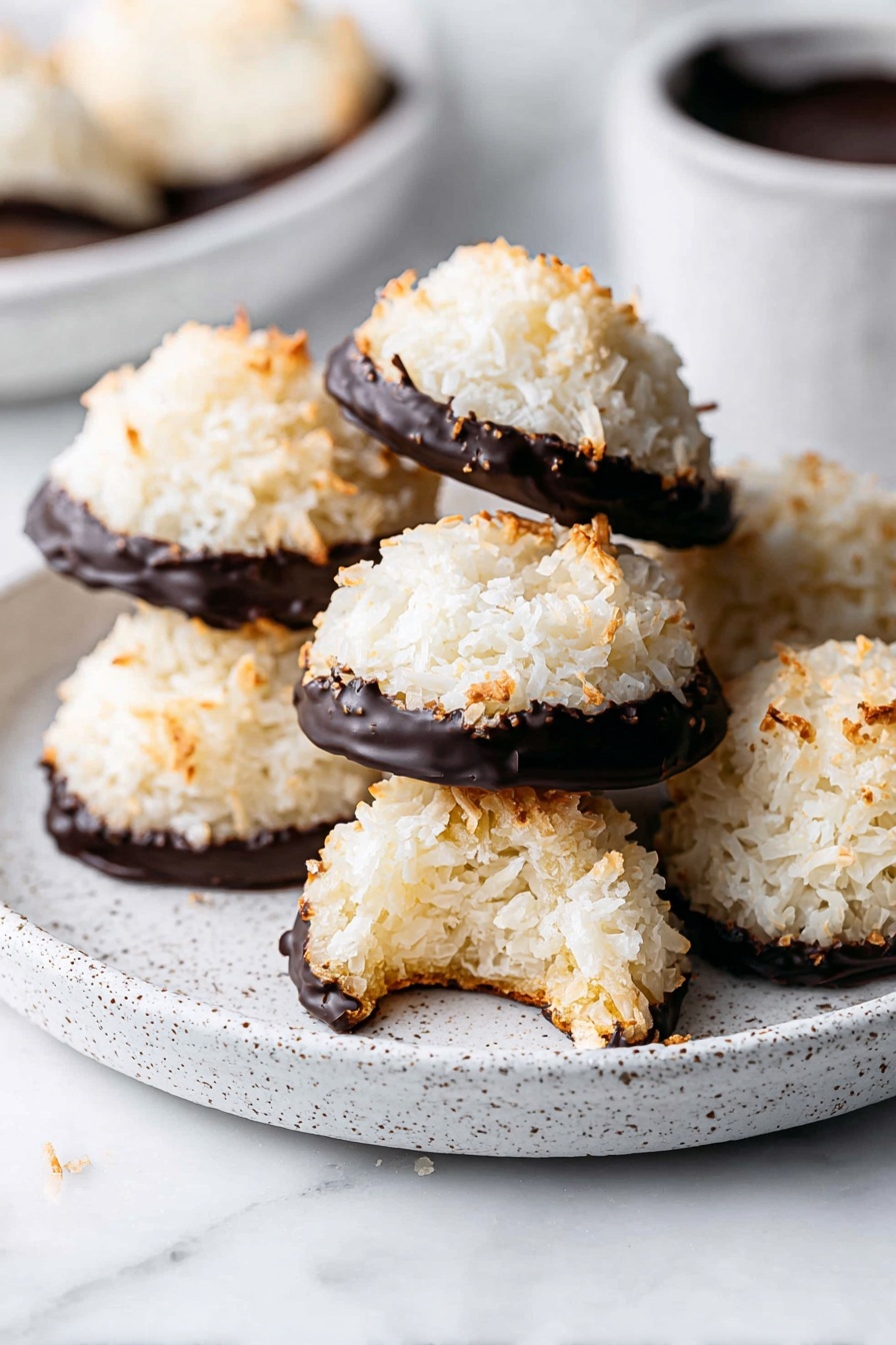 The image shows several round coconut macaroons with a rough, white, and slightly toasted top layer. Each macaroon has a rich, dark chocolate coating covering the bottom half, creating a sharp color contrast. They are arranged on a speckled white plate placed on a white marbled surface. One macaroon in the front has a bite taken out of it, revealing a soft, fibrous inside. In the background, there is a white bowl with dark chocolate sauce, blurred slightly to keep focus on the macaroons. photo taken with an iphone --ar 2:3 --v 7 - German Coconut Macaroons, coconut macaroon recipe, tropical cookies, easy coconut cookies, kokosmakronen