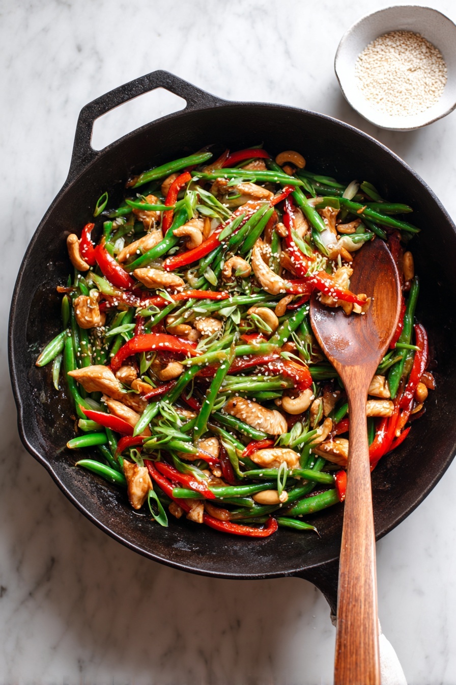 The image shows a black cast iron pan filled with a colorful stir-fry. The stir-fry has about four layers: large light brown pieces of cooked chicken evenly spread; bright green string beans scattered throughout; thin, long red bell pepper strips mixed in; and small slices of green onions on top. There are also pale cashew nuts adding texture and white sesame seeds sprinkled over everything. A wooden spoon rests inside the pan on the right side, slightly covered by some food. The pan is placed on a white marbled surface with a small white bowl of sesame seeds nearby. Photo taken with an iphone --ar 2:3 --v 7 - Healthy Orange Chicken Stir Fry with Cauliflower Rice, orange chicken stir fry healthy, low carb cauliflower rice recipes, healthy chicken stir fry, citrus chicken dinner