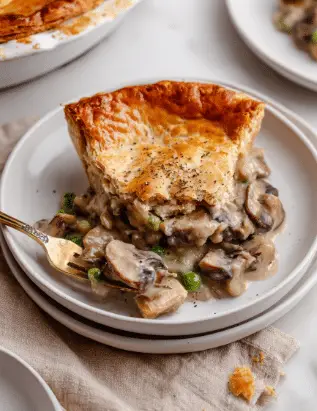 A white round plate holds a portion of vegan mushroom pot pie with two clear layers: the bottom layer is a creamy mushroom filling with sliced brown mushrooms, green peas, and a thick sauce, while the top layer is a golden-brown flaky crust slightly cracked with a sprinkled touch of black pepper. A bronze fork on the plate lifts a piece of the mushroom filling, showing its chunky texture. The plate sits on a beige cloth with a small part of a white marbled surface visible around it. In the background, part of a large round pot pie with a well-baked golden crust is visible on the left, and a white plate with another piece of pie shows on the right. Photo taken with an iphone --ar 2:3 --v 7 - Vegan Mushroom Pot Pie, vegan savory pie, plant-based mushroom pie, dairy-free mushroom pot pie, vegan comfort food