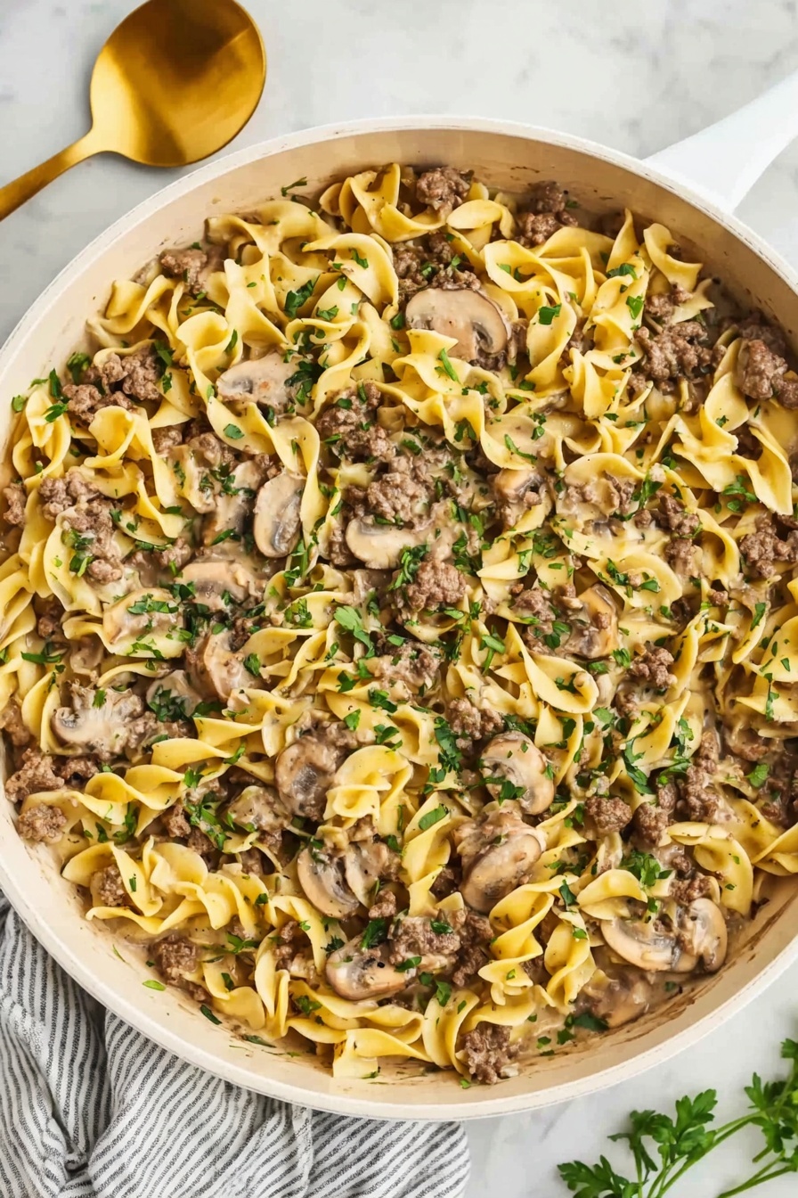 The image shows a wide white pan filled with creamy egg noodles mixed with sliced mushrooms and small pieces of cooked ground beef. The noodles are soft yellow with a smooth texture, spread evenly across the pan. The mushrooms, pale brown and slightly shiny, are mixed throughout the noodles. The beef is browned and scattered on top and inside the noodles in small chunks. Fresh green herbs are sprinkled over the whole dish, adding a pop of color. The pan sits on a white marble surface, next to a gold spoon and white cloth with gray stripes. Photo taken with an iphone --ar 2:3 --v 7 - Ground Beef Stroganoff, One-Pot Ground Beef Stroganoff, Easy Beef Stroganoff, Creamy Beef Pasta, Quick Weeknight Dinner