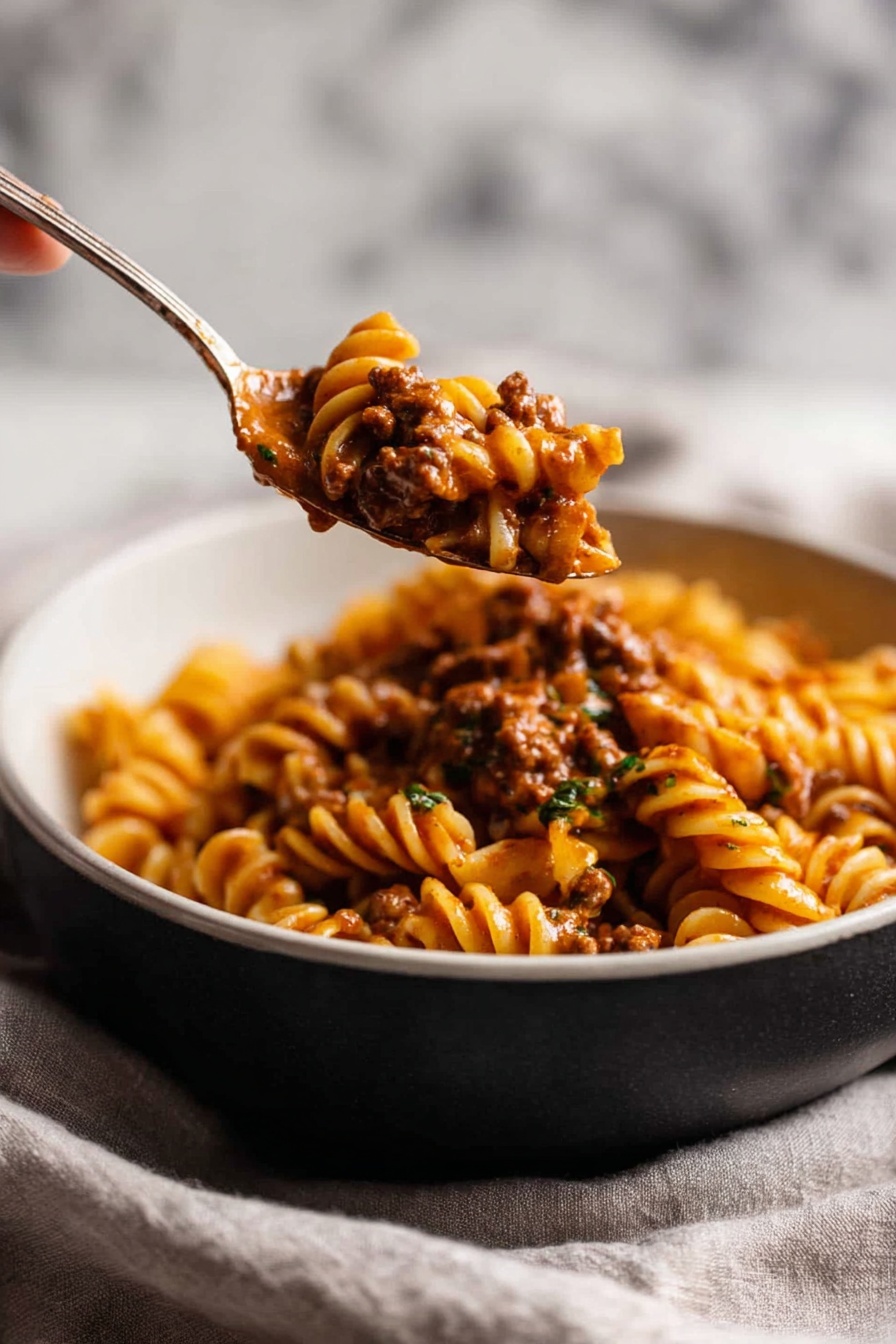 A close-up view shows spiral pasta mixed with a thick, rich brown sauce that includes small pieces of ground meat, all held on a spoon lifting from a two-layer bowl with a dark top half and a white bottom half. The pasta is covered fully by the sauce, which looks creamy and dense, with some green herb bits scattered inside. The bowl sits on a soft light gray cloth with a blurred white marbled texture in the background. A woman's hand holds the spoon from the left side, drawing the pasta up. photo taken with an iphone --ar 2:3 --v 7 - Creamy Tomato Beef Pasta, creamy tomato beef pasta, beef pasta recipe, one-pot beef pasta, easy beef pasta