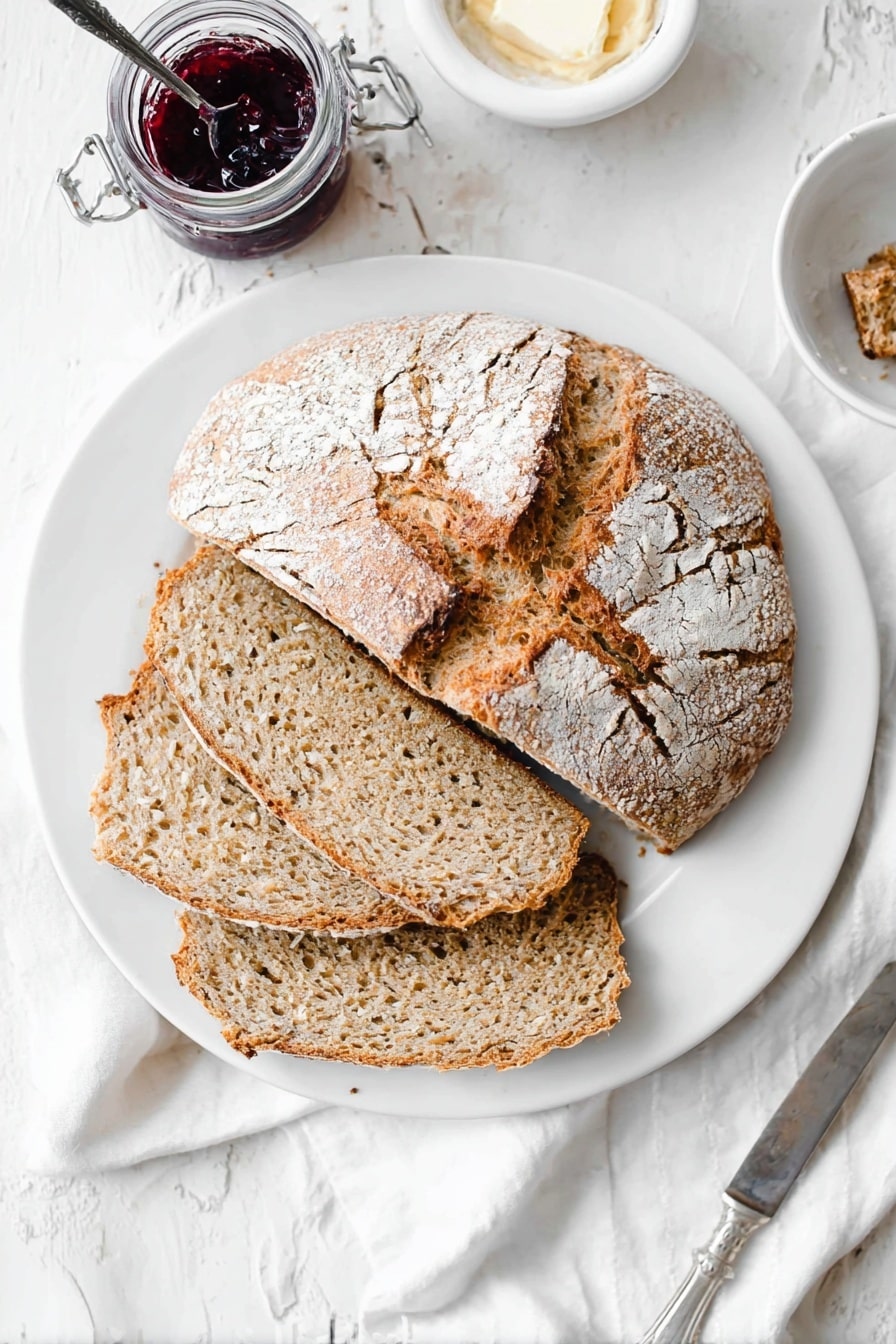 A white plate sits on a white marbled textured surface, holding a round loaf of rustic bread that is partially sliced into four thick pieces. The top half of the loaf has a golden-brown crust with a dusting of white flour and a rough, cracked texture. The inside of the bread is light brown with a soft, porous texture. To the right of the plate, a small jar of dark red fruit jam with a small spoon inside is visible, along with a small white dish containing a pale spread. A silver knife rests on a white cloth napkin beside the plate. Photo taken with an iphone --ar 2:3 --v 7 - Irish Soda Bread, Easy Irish Soda Bread, homemade Irish bread, quick Irish bread, simple soda bread