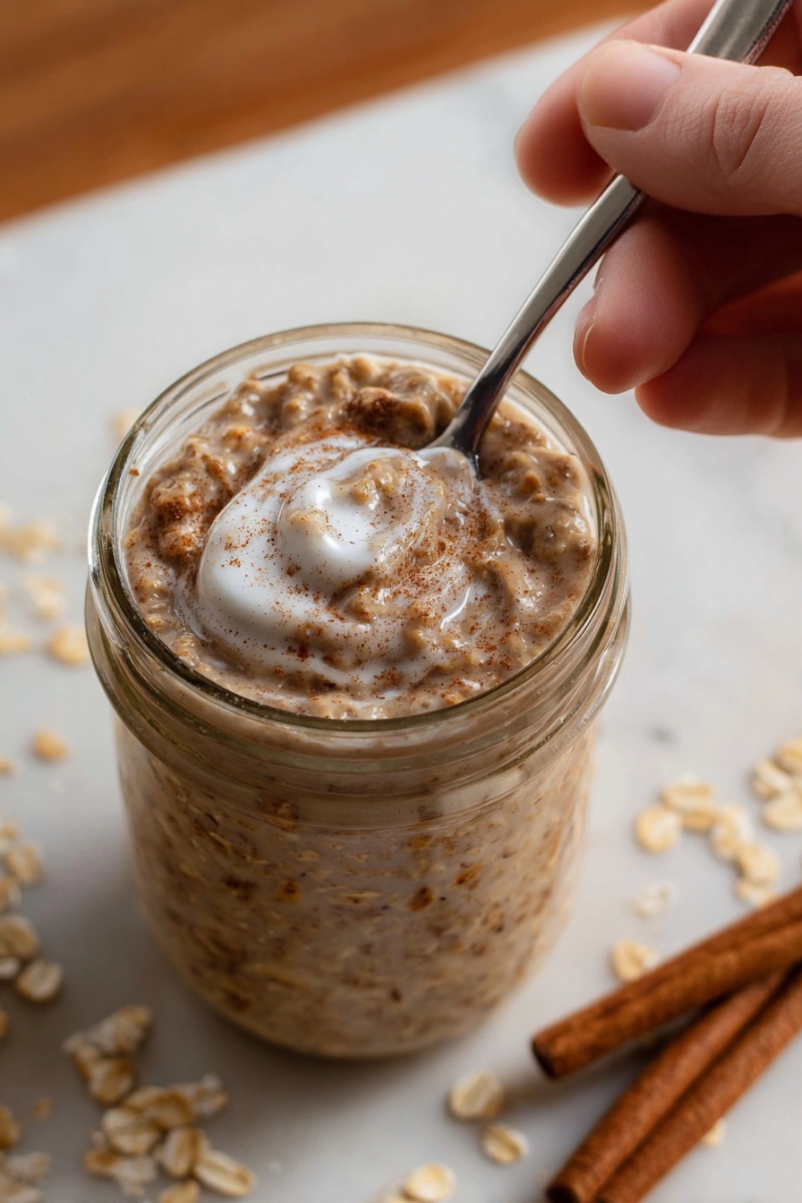 A glass jar is filled with a creamy oatmeal mixture that has a light brown color with visible oats and specks of cinnamon mixed throughout. The top layer shows a soft texture with a swirl of white milk or cream blending in, giving it a slightly glossy look. A metal spoon is dipped into the jar from the top right, held by a woman's hand. The background is a white marbled texture with scattered oats and two cinnamon sticks placed near the jar. The overall scene has warm tones emphasizing the cozy feel of the oatmeal. photo taken with an iphone --ar 2:3 --v 7 - Cinnamon Roll Overnight Oats, overnight oats with cinnamon, easy healthy breakfast, make-ahead breakfast recipes, nutritious morning ideas