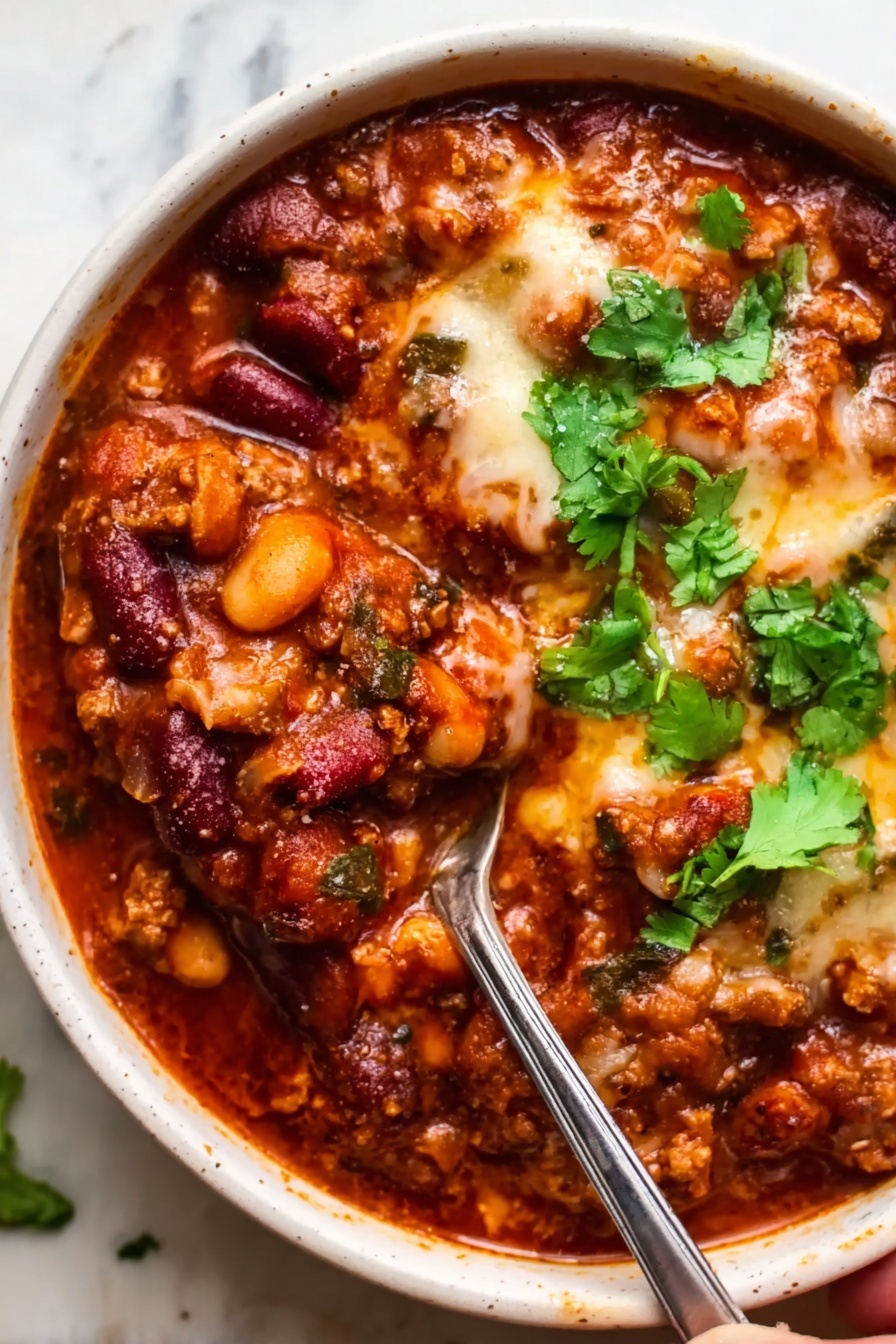 A white bowl filled with a thick chili, showing layers of rich red tomato sauce, dark red kidney beans, beige beans, and cooked ground meat mixed together. Melted cheese with a slightly browned top covers part of the chili, adding a creamy texture. Fresh green cilantro leaves sit on top, adding a bright pop of color. A silver spoon is inside the bowl, and a woman's hand is holding the spoon. The background is a white marbled surface. photo taken with an iphone --ar 2:3 --v 7 - Hearty Beef Chili, easy beef chili recipe, hearty chili dinner, comforting beef chili, flavorful chili ideas