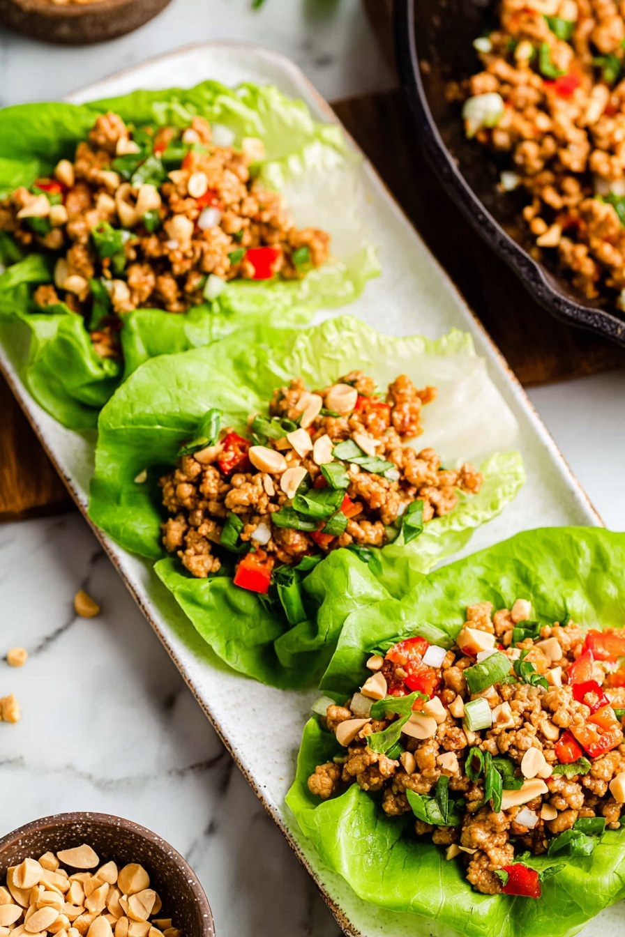 Three lettuce wraps are placed side by side on a long white plate on a white marbled surface. Each wrap has a base layer of bright green lettuce leaves that are smooth and slightly curled at the edges. On top, there is a thick layer of light brown cooked meat mixed with small diced red and white vegetables and green herbs. The wraps are sprinkled with chopped peanuts, adding a light beige color and a rough texture on top. In the background, there is a black skillet with more of the cooked meat mixture and a small bowl with scattered chopped peanuts. photo taken with an iphone --ar 2:3 --v 7 - Asian Chicken Lettuce Wraps, Asian Chicken Lettuce Wraps recipe, Healthy Chicken Lettuce Wraps, Easy Asian Lettuce Wraps, Quick Dinner Lettuce Wraps