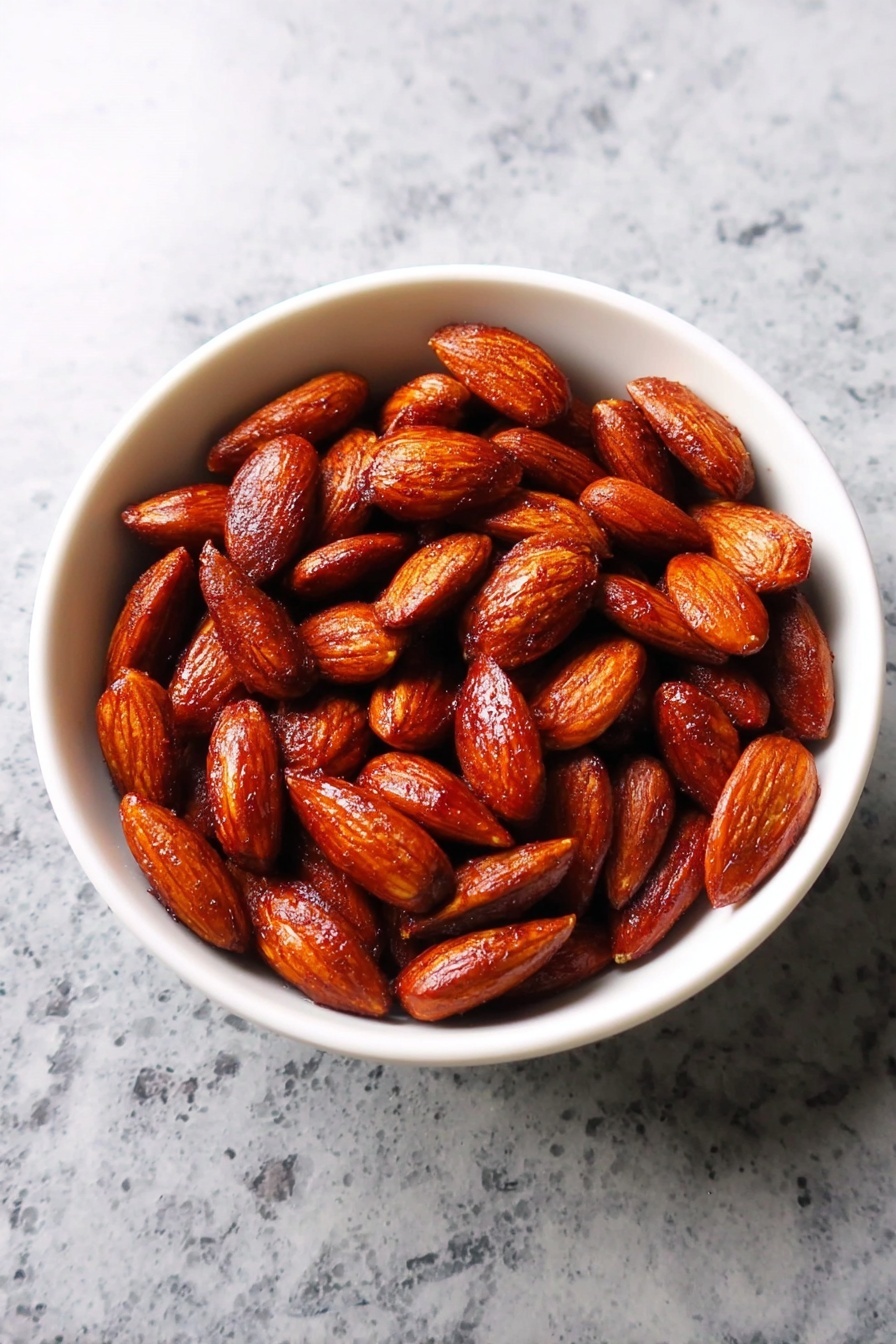 A white bowl filled with a single layer of roasted almonds that have a shiny, dark reddish-brown color and a slightly oily texture, sitting on a white marbled surface. The almonds are whole and look crispy with a rich roasted finish. Photo taken with an iphone --ar 2:3 --v 7 - Spicy Roasted Almonds, Spicy Roasted Almonds recipe, Crunchy spicy nuts, Easy snack recipes, Homemade seasoned almonds