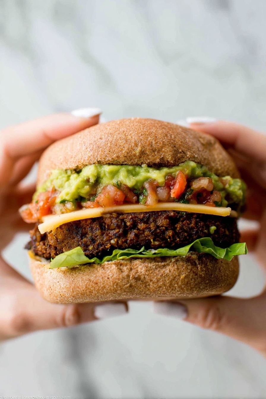 A close-up image of a sandwich held by two woman's hands with light-colored nail polish, showing four layers inside a soft, brown whole wheat bun. The bottom layer is green leaf lettuce, followed by a thick, dark brown veggie patty with a coarse, chunky texture. On top of the patty is a slice of pale yellow cheese partially melted, then a layer of chunky green guacamole with a creamy texture partly mixed with bright red tomato salsa. The top bun is slightly squished down over the layers. The background features a white marbled texture. photo taken with an iphone --ar 2:3 --v 7 - Best Black Bean Burgers, vegetarian burger recipes, easy black bean burgers, healthy burger ideas, flavorful veggie burgers