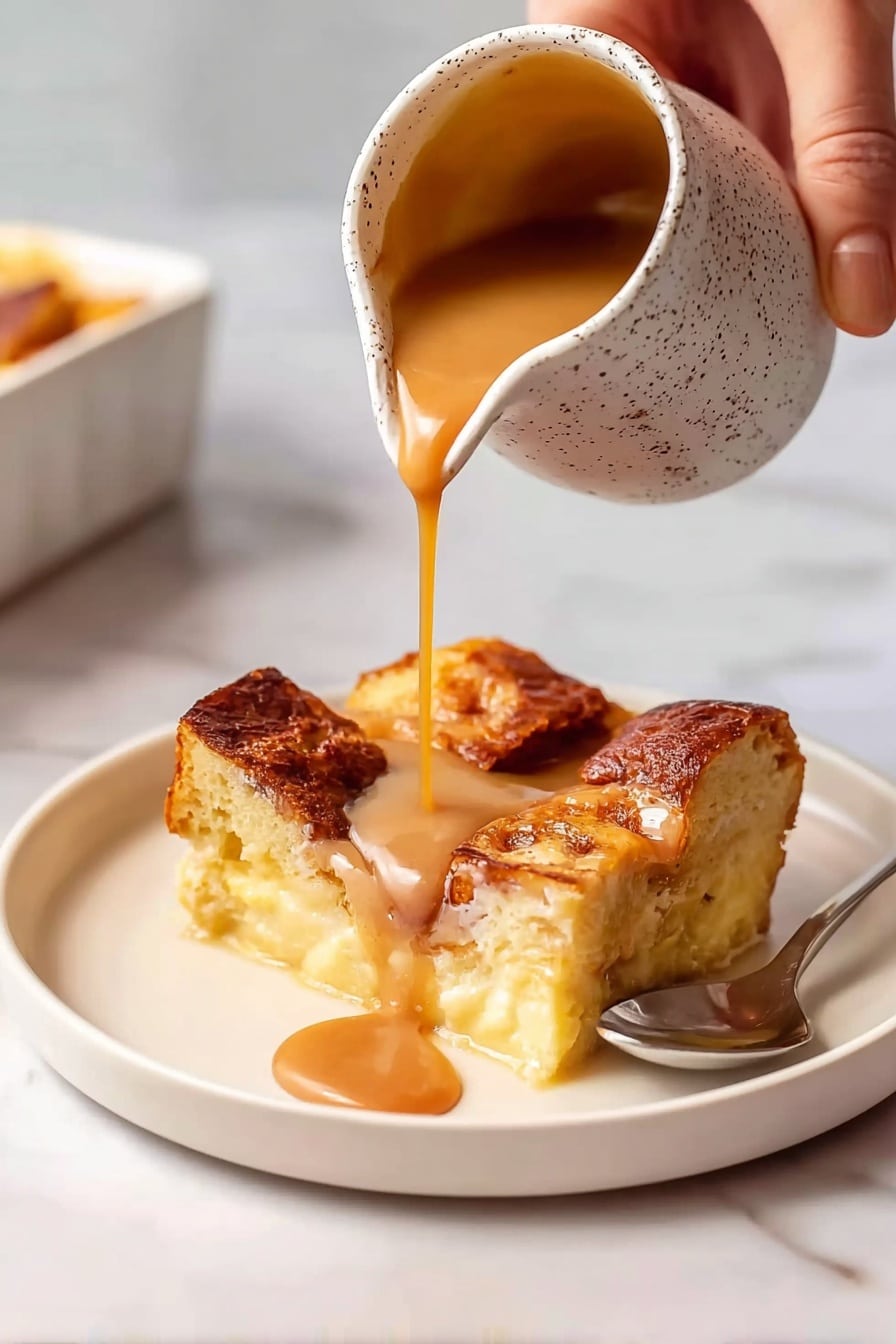 A white round plate holds a square piece of bread pudding with four large, golden brown, crisp bread chunks on top, showing a soft, creamy, pale yellow inside with visible texture from baked egg custard and bread soaked within. A woman's hand is pouring a smooth, thick caramel sauce of warm light brown color from a small, speckled, white jug onto the bread pudding, with the sauce flowing in a steady stream and pooling over the top, enhancing its shine. A spoon rests on the plate beside the dessert, and the whole scene is set on a white marbled surface. photo taken with an iphone --ar 2:3 --v 7 - Caramel Bread Pudding with Homemade Sauce, caramel bread pudding, homemade caramel sauce recipe, comforting dessert ideas, easy bread pudding dessert