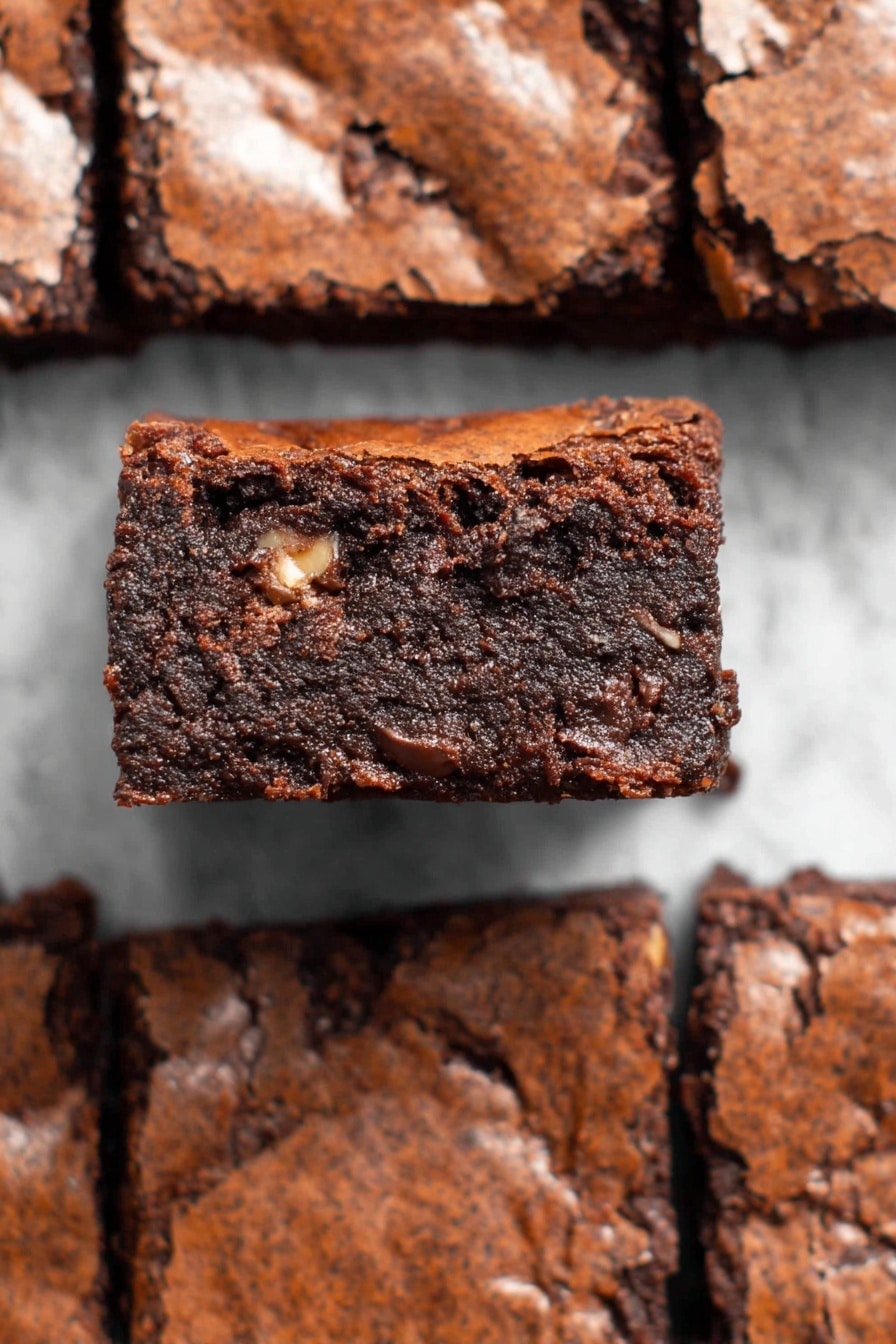 A square chocolate brownie is lifted above others around it, showing its thick, dark brown interior with a slightly shiny and cracked top layer. The inside looks dense and moist with some lighter brown pieces embedded in it, suggesting nuts or chocolate chunks. The other brownies beneath form a grid pattern on a white marbled texture. The lighting highlights the brownie’s rich texture and soft crumb edge. photo taken with an iphone --ar 2:3 --v 7 - Fudgy Almond Flour Brownies, healthy chocolate brownies, gluten-free dessert recipes, easy almond flour brownies, indulgent chocolate treats