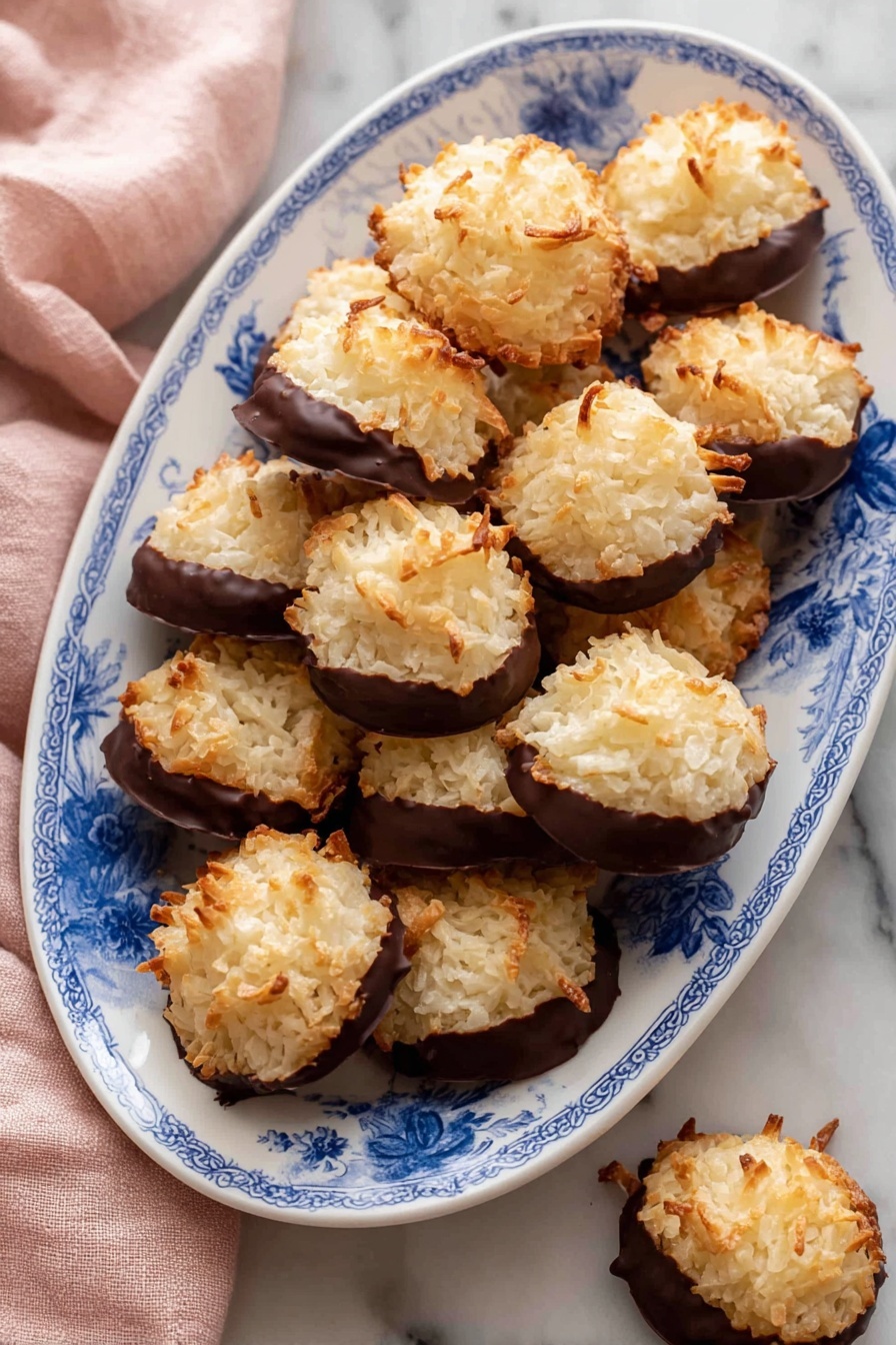 The image shows a white oval plate with a blue floral pattern filled with round coconut cookies. Each cookie has two layers: the top layer is golden brown with a rough, shredded texture of toasted coconut, and the bottom layer is dipped in smooth, dark chocolate that covers about half the cookie. The cookies are piled on the plate over a white marbled surface, with a soft pink cloth partially visible on the left side. One cookie is set just outside the plate at the bottom right. The photo taken with an iphone --ar 2:3 --v 7 - Delicious Coconut Macaroons with Chocolate, coconut macaroons, chocolate-covered macaroons, easy coconut treats, chewy coconut cookies
