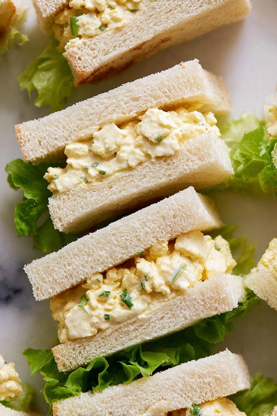 The image shows small sandwich pieces with two layers of soft, white bread surrounding a creamy, pale yellow egg salad filling mixed with small bits of egg white and sprinkled with green herbs. The sandwiches are placed on top of bright green lettuce leaves, which add a fresh contrast to the light-colored sandwiches. The texture of the bread looks soft and slightly fluffy, while the filling appears smooth but textured with tiny egg pieces. The background surface is a white marbled texture. photo taken with an iphone --ar 2:3 --v 7 - Creamy Soft Boiled Egg Sandwich, egg sandwich recipe, soft boiled egg breakfast, luxurious egg sandwich, easy egg sandwich idea