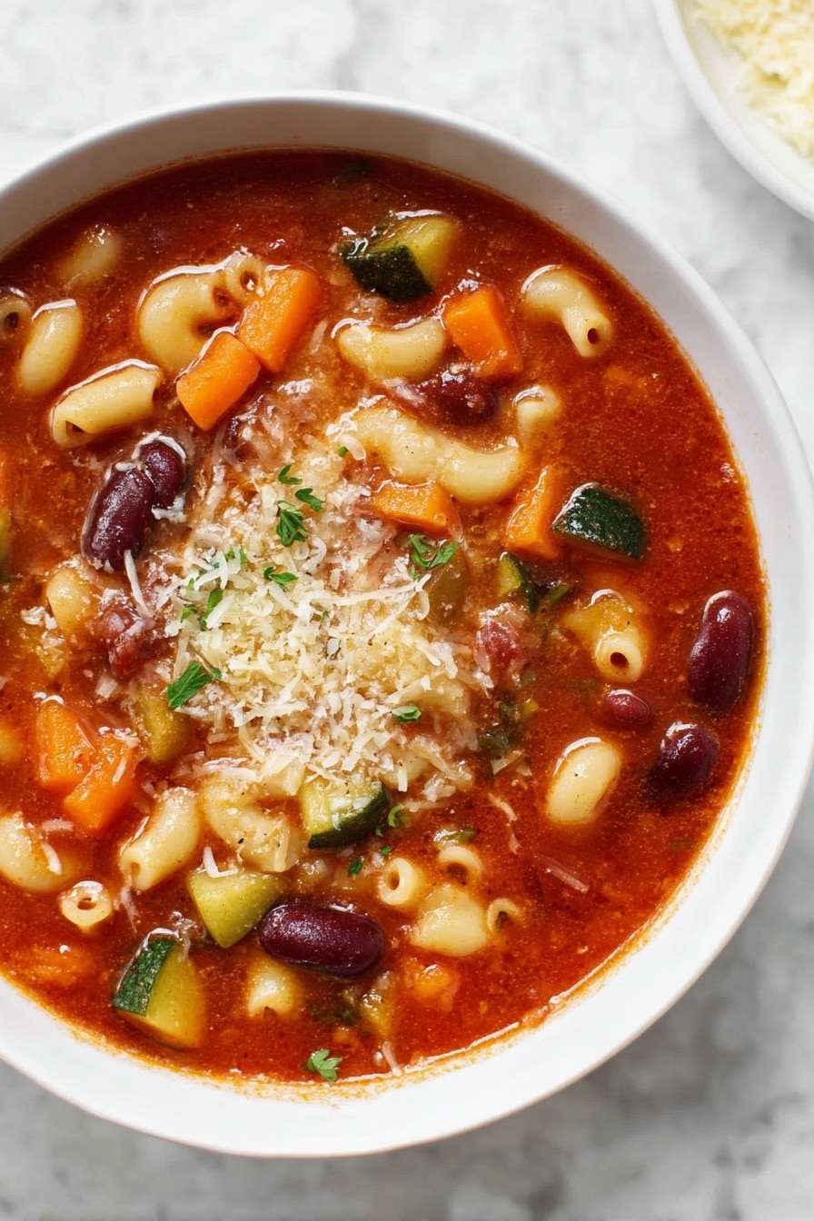A white bowl filled with a thick tomato-based soup showing three main layers: a deep red-orange broth as the base with a shiny, smooth texture; the middle layer consists of mixed ingredients including curved pale yellow elbow pasta, dark red kidney beans, orange carrot slices, and green zucchini and green beans; the top layer is sprinkled with grated light brown cheese and small bits of green herbs, scattered evenly. The bowl sits on a white marbled surface. Photo taken with an iphone --ar 2:3 --v 7 - Instant Pot Minestrone Soup, easy minestrone soup, vegetarian Instant Pot soup, healthy one-pot soup, quick vegetable soup