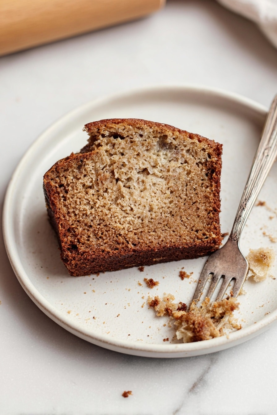 A single piece of brown banana bread with a slightly darker crust sits on a white plate. The bread slice has a soft, spongy texture with small holes and bits inside. A silver fork rests on the plate, holding a small bite of the bread. Crumbs are scattered around the plate, which is placed on a white marbled surface near a wooden rolling pin. Photo taken with an iphone --ar 2:3 --v 7 - Best Banana Bread with Cinnamon Crunch, moist banana bread with cinnamon topping, easy banana bread recipe, delicious banana loaf with crunch, overripe banana bread ideas