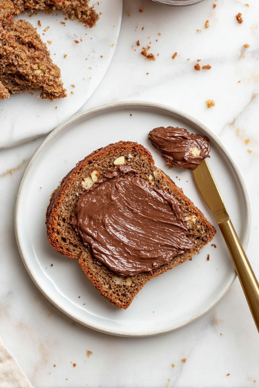 A white plate with one thick slice of brown bread that has a rough, textured surface and visible small nuts inside. The top is spread with a thick, shiny layer of smooth chocolate spread. Near the bread, there is a smaller piece with chocolate spread on it. A gold knife with chocolate spread on its blade rests on the plate. The background is a white marbled surface with small bread crumbs scattered around. Photo taken with an iphone --ar 2:3 --v 7 - Best Banana Bread with Cinnamon Crunch, moist banana bread with cinnamon topping, easy banana bread recipe, delicious banana loaf with crunch, overripe banana bread ideas