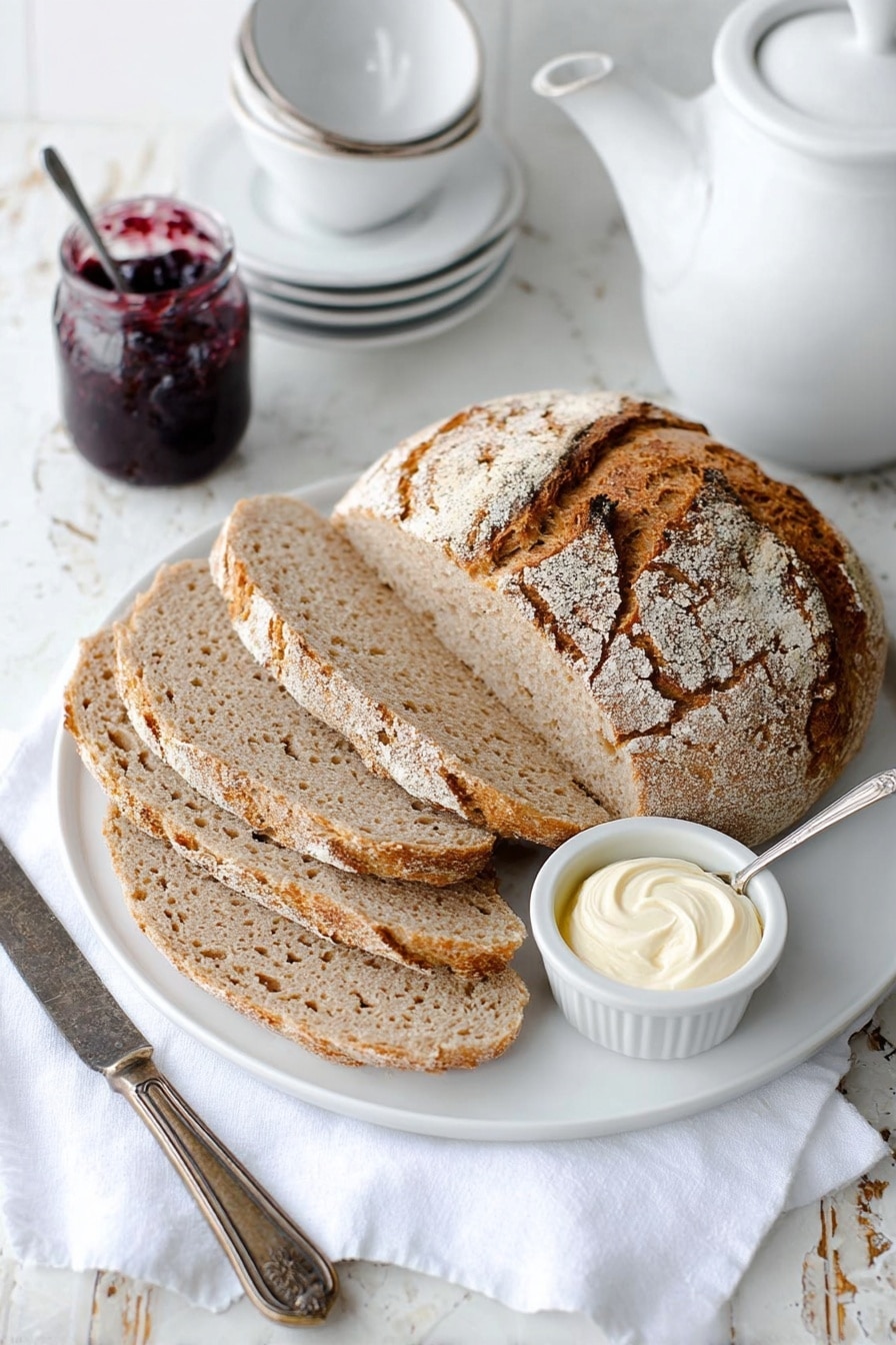 A white round plate holds a loaf of rustic bread with a cracked, golden-brown crust dusted lightly with flour. Four thick slices of the bread lie fanned out in front of the loaf, showing a soft, light brown interior with a slightly coarse texture. To the right of the bread, there is a small white dish with a swirl of creamy white butter and a small glass jar filled with dark purple jam, which has a silver spoon resting inside. A vintage metal knife lies on a white cloth napkin beside the plate. The background is a white marbled texture with a white teapot and stacked white teacups placed nearby. Photo taken with an iphone --ar 2:3 --v 7 - Irish Soda Bread, Easy Irish Soda Bread, homemade Irish bread, quick Irish bread, simple soda bread