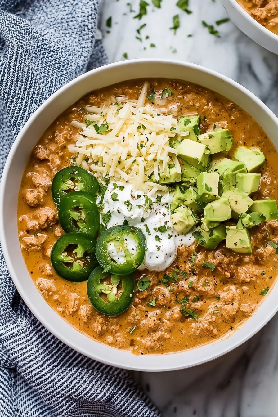 A white bowl filled with a thick, creamy, orange-brown base layer that looks like chili or stew with bits of meat. On top, there are four distinct sections: thin green jalapeño slices on the left, shredded pale yellow cheese below, cubed light green avocado pieces to the right, and a dollop of white sour cream in the center. The toppings are sprinkled with chopped green herbs and some black pepper. The bowl sits on a white marbled surface with a blue and white striped cloth partly under it. Photo taken with an iphone --ar 2:3 --v 7 - Creamy White Bean Turkey Chili, hearty turkey chili with white beans, healthy white bean turkey chili, comforting turkey chili recipes, easy white bean chili