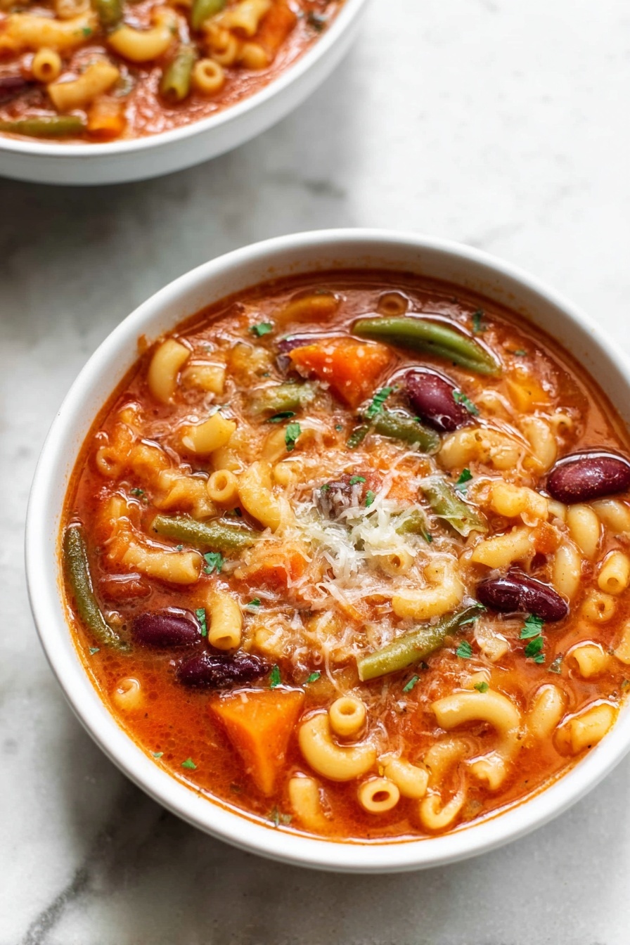 A white bowl filled with a rich, orange-red tomato soup, layered with elbow macaroni pasta, dark red kidney beans, sliced orange carrots, and green beans. The soup is topped with a sprinkling of finely grated cheese that slightly melts into the warm broth, and small green herb pieces are scattered on top. The bowl sits on a white marbled surface. In the background, another white bowl of the same soup is partially visible. photo taken with an iphone --ar 2:3 --v 7 - Instant Pot Minestrone Soup, easy minestrone soup, vegetarian Instant Pot soup, healthy one-pot soup, quick vegetable soup