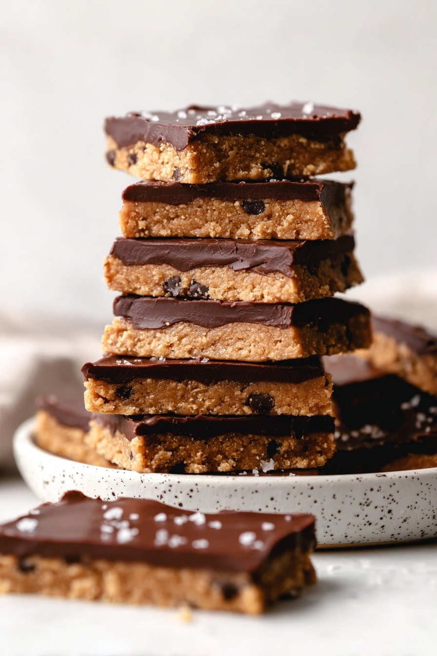 The image shows several rectangular dessert bars with two main layers. The bottom layer is thick, light golden brown with a crumbly texture that looks like a cookie or biscuit base. The top layer is a smooth, dark chocolate layer with a shiny surface. Large flakes of sea salt and small dark bits are sprinkled over the chocolate, adding texture and contrast. The bars are arranged closely on a white marbled surface, with some bars overlapping slightly. photo taken with an iphone --ar 2:3 --v 7 - Healthy Protein Cookie Dough Bark, healthy protein snacks, guilt-free cookie dough treats, quick healthy dessert, high-protein sweet bites