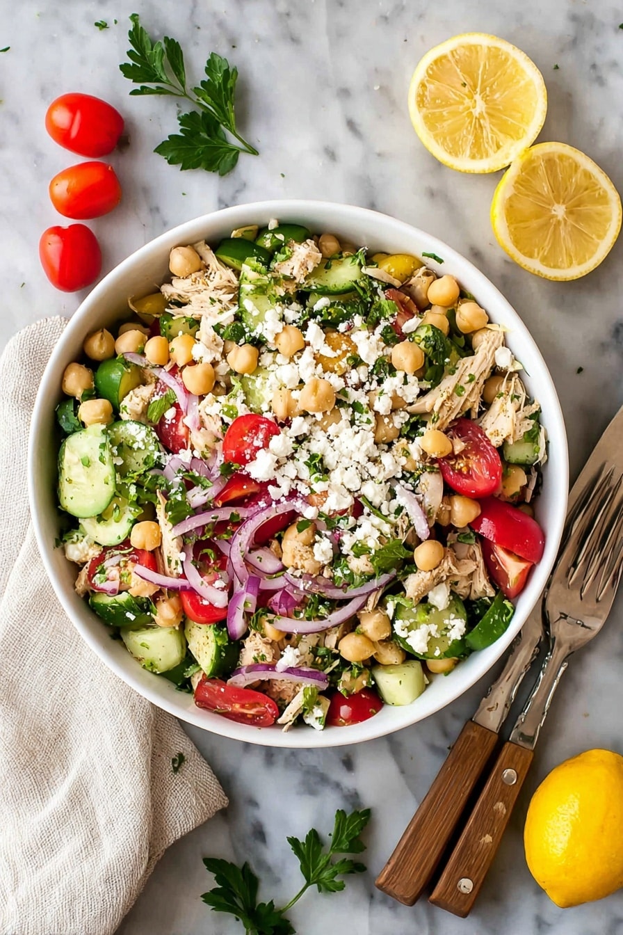 A white bowl filled with a colorful salad sits on a white marbled surface. The salad has many layers: at the base, there are green cucumber slices mixed with bright red grape tomatoes, beige chickpeas scattered throughout, and small pieces of light brown cooked chicken. Thin, curved strips of purple onion are spread inside, along with chopped green herbs. Crumbled white cheese is sprinkled on top, adding a crumbly texture. Around the bowl, there are whole grape tomatoes, parsley leaves, and two lemon wedges with a bright yellow rind. On the right side, a white cloth holds a fork and knife with wooden handles. Photo taken with an iphone --ar 2:3 --v 7 - Healthy Chickpea Tuna Salad with Fresh Veggies, healthy tuna salad, quick nutritious lunch, easy chickpea tuna recipe, fresh veggie salad