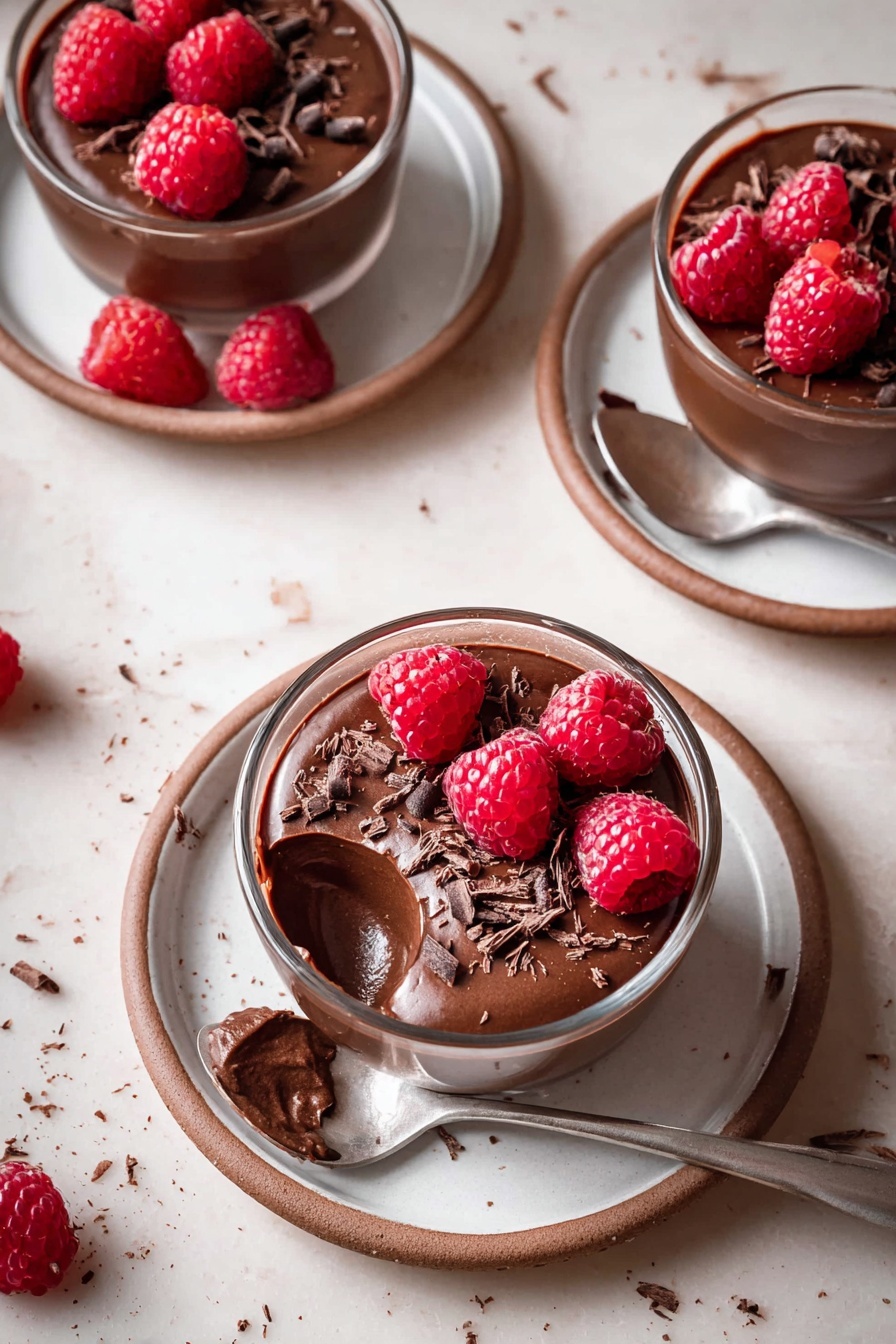 The image shows three small clear glass cups filled with smooth dark brown chocolate mousse, each one topped with bright red fresh raspberries and small dark chocolate shavings. The mousse inside the cups looks creamy with a shiny surface, and a spoon with a scoop of mousse rests on one of the white plates holding a cup. Each cup is placed on a white plate with a light brown rim, set on a white marbled surface that has scattered chocolate shavings and a few raspberries around. The lighting highlights the rich colors and textures, giving the mousse a soft, inviting look. Photo taken with an iphone --ar 2:3 --v 7 - Vegan Chocolate Mousse with Raspberry Compote, vegan chocolate mousse, dairy-free chocolate mousse, plant-based dessert, raspberry compote recipe