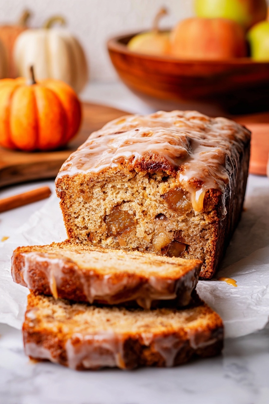 The image shows a loaf of bread with two slices cut and placed in front. The bread is golden brown with a textured crust, and the top layer is covered with a shiny glaze that drips slightly down the sides. Inside, the bread looks soft and moist, with visible chunks of fruit or nuts. The bread rests on white parchment paper over a white marbled surface. In the background, there are blurred autumn-themed items like small pumpkins and a wooden bowl with apples. Photo taken with an iphone --ar 2:3 --v 7 - Amish Apple Fritter Bread, apple fritter bread with glaze, cinnamon apple bread, easy apple bread recipe, sweet apple bread dessert