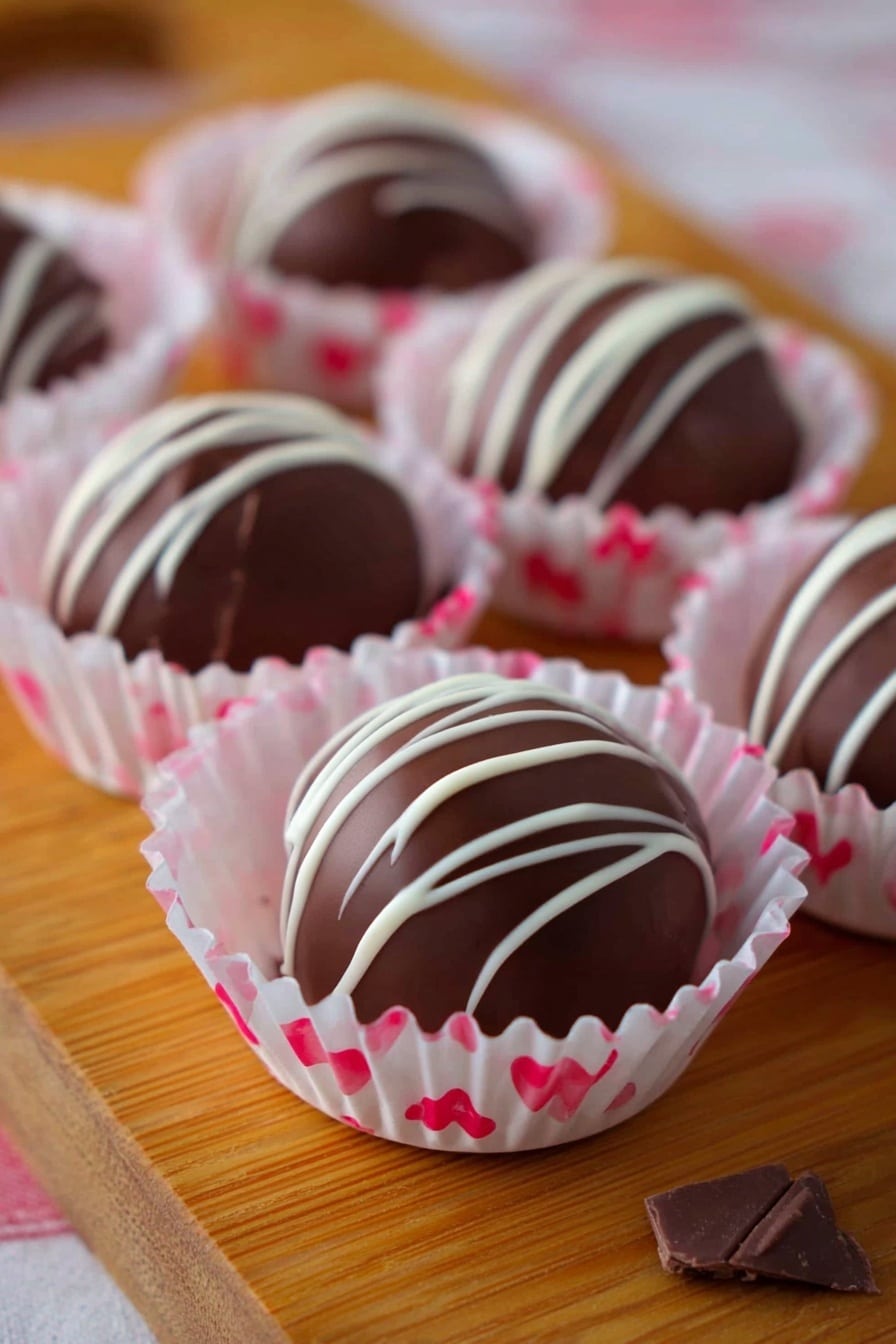 This image shows a wooden board with smooth round dark chocolate truffles placed in white paper cups with pink and red hearts design, each truffle drizzled with thin white chocolate lines on top in irregular patterns. The truffles have a shiny dark brown surface and sit in a single layer in their cups. The soft pink color of the paper cups contrasts gently with the rich chocolate, making each piece look delicate and inviting. Small pieces of chocolate can be seen around the cups. The photo taken with an iphone --ar 2:3 --v 7 - Strawberry White Chocolate Truffles, strawberry dessert recipes, easy fruit truffles, white chocolate confections, homemade chocolate truffles