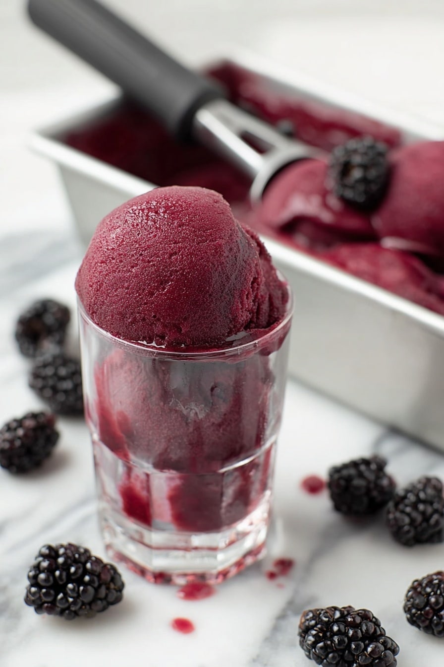 A clear glass holds one large scoop of deep purple blackberry sorbet with a smooth, slightly icy texture, filling the top of the glass and dripping slightly down the sides. Behind it, there is a white metal container filled with more of the same sorbet, some blackberries resting on top, with a black handled ice cream scoop inside. Around the glass on a white marbled surface, several plump blackberries are scattered casually. The scene is bright and fresh-looking, focusing on the rich color and texture of the sorbet. photo taken with an iphone --ar 2:3 --v 7 - Blackberry Sorbet, Blackberry Sorbet recipe, healthy fruit sorbet, easy homemade sorbet, vegan berry dessert