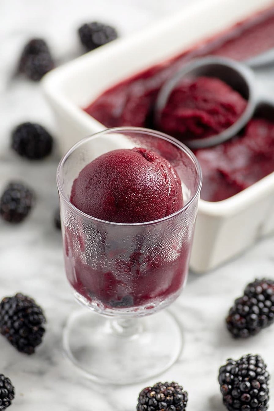 A clear glass holds a single scoop of dark purple-blackberry sorbet, showing a smooth and slightly shiny texture with a few light reflections. The sorbet is rounded on top and fills the glass closely, with some frost visible on the outside of the glass, which stands on a white marbled surface. Surrounding the glass are several fresh blackberries scattered around. Behind the glass is a white rectangular tray filled with more of the same dark purple sorbet, with an ice cream scoop resting inside the tray. The background and all surfaces have a white marbled texture. photo taken with an iphone --ar 2:3 --v 7 - Blackberry Sorbet, Blackberry Sorbet recipe, healthy fruit sorbet, easy homemade sorbet, vegan berry dessert