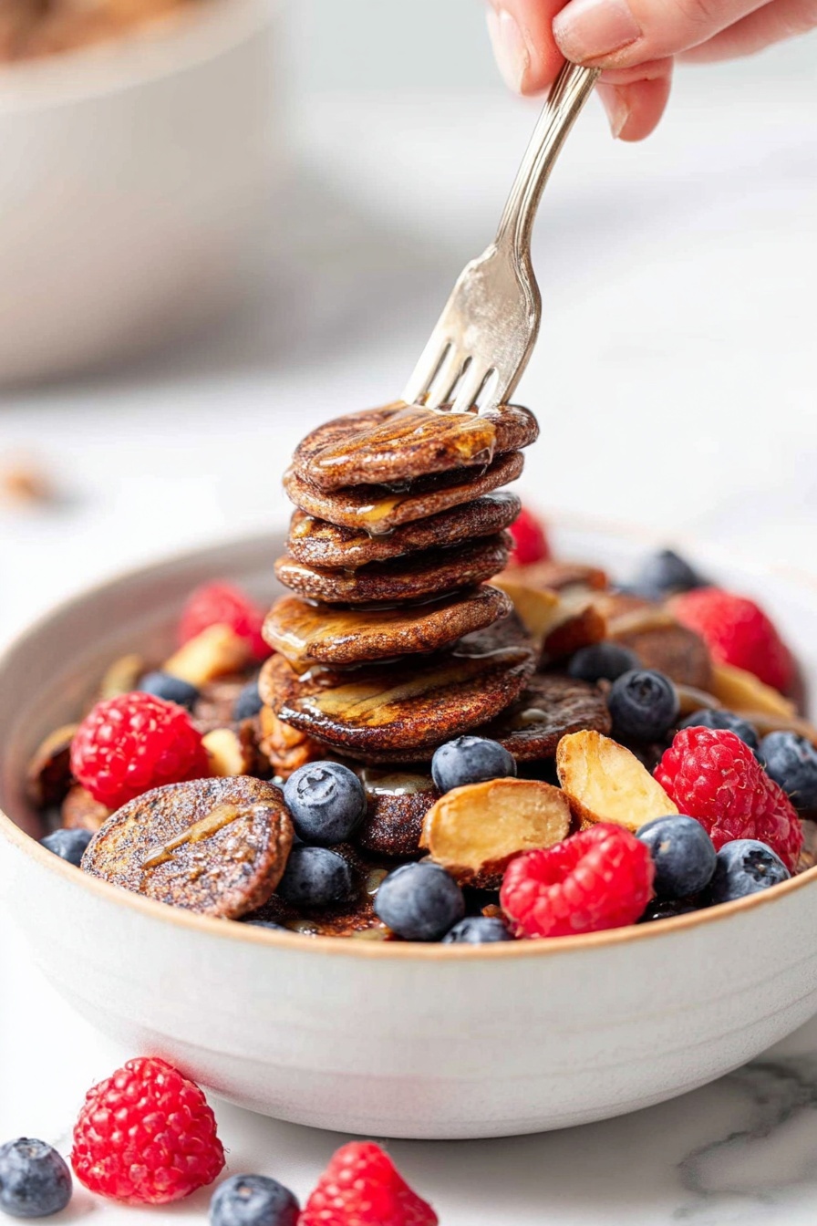 A white bowl filled with many small, dark brown mini pancakes roughly stacked and topped with a few drizzles of light brown syrup. Fresh blueberries and bright red raspberries are mixed throughout the bowl, with some raspberries placed around the bowl on a white marbled surface. A woman's hand holding a fork lifts a stack of five mini pancakes from the bowl. The background is bright and blurred, focusing on the bowl. Photo taken with an iphone --ar 2:3 --v 7 - Mini Pancake Cereal Bowl with Berries, pancake cereal recipes, adorable breakfast ideas, healthy pancake breakfast, berry breakfast bowls