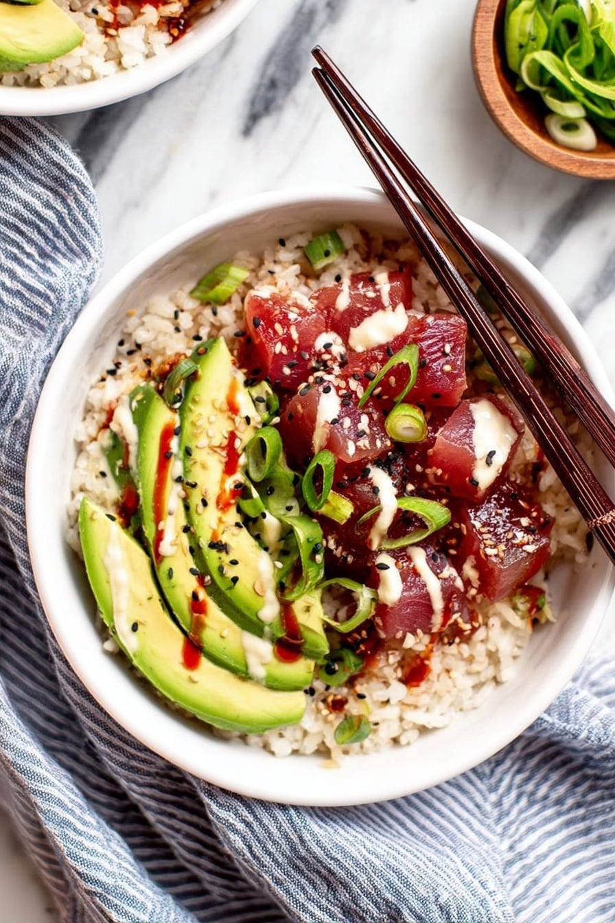 A white bowl filled with three main layers: the bottom layer is light brown cooked rice, the middle layer shows bright green avocado slices arranged in a fan shape on one side, and the top layer is deep red cubed raw fish covered with light cream sauce and red sauce drizzles. Small black sesame seeds and sliced green onions are scattered over the avocado and fish. Two dark wooden chopsticks rest diagonally across the bowl on top of the food. The bowl is placed on a white marbled surface with a blue striped cloth beside it, and a small wooden bowl with green sliced vegetables is partially visible in the background. Photo taken with an iphone --ar 2:3 --v 7 - Spicy Tuna Poke Bowl, tuna poke bowl recipe, spicy seafood bowl, quick healthy poke, homemade poke bowl