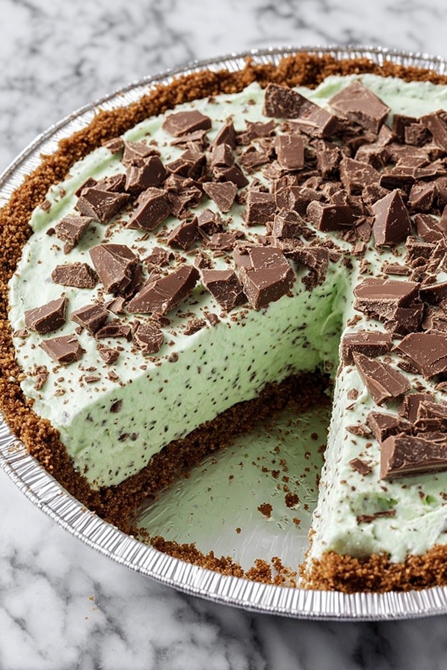 The image shows a pie with three main layers. The bottom layer is a crumbly brown crust that fills the pie tin. The middle layer is thick and pale green with small dark specks mixed in, giving it a soft, creamy texture. The top layer is made up of broken pieces of milk chocolate scattered across the green filling. One slice has been removed, showing the inside structure clearly. The pie tin is silver and the background is a white marbled surface. Photo taken with an iphone --ar 2:3 --v 7 - Mint Chocolate Pie, no-bake mint chocolate dessert, easy mint chocolate pie recipe, quick chocolate pie with mint, best mint chocolate pie