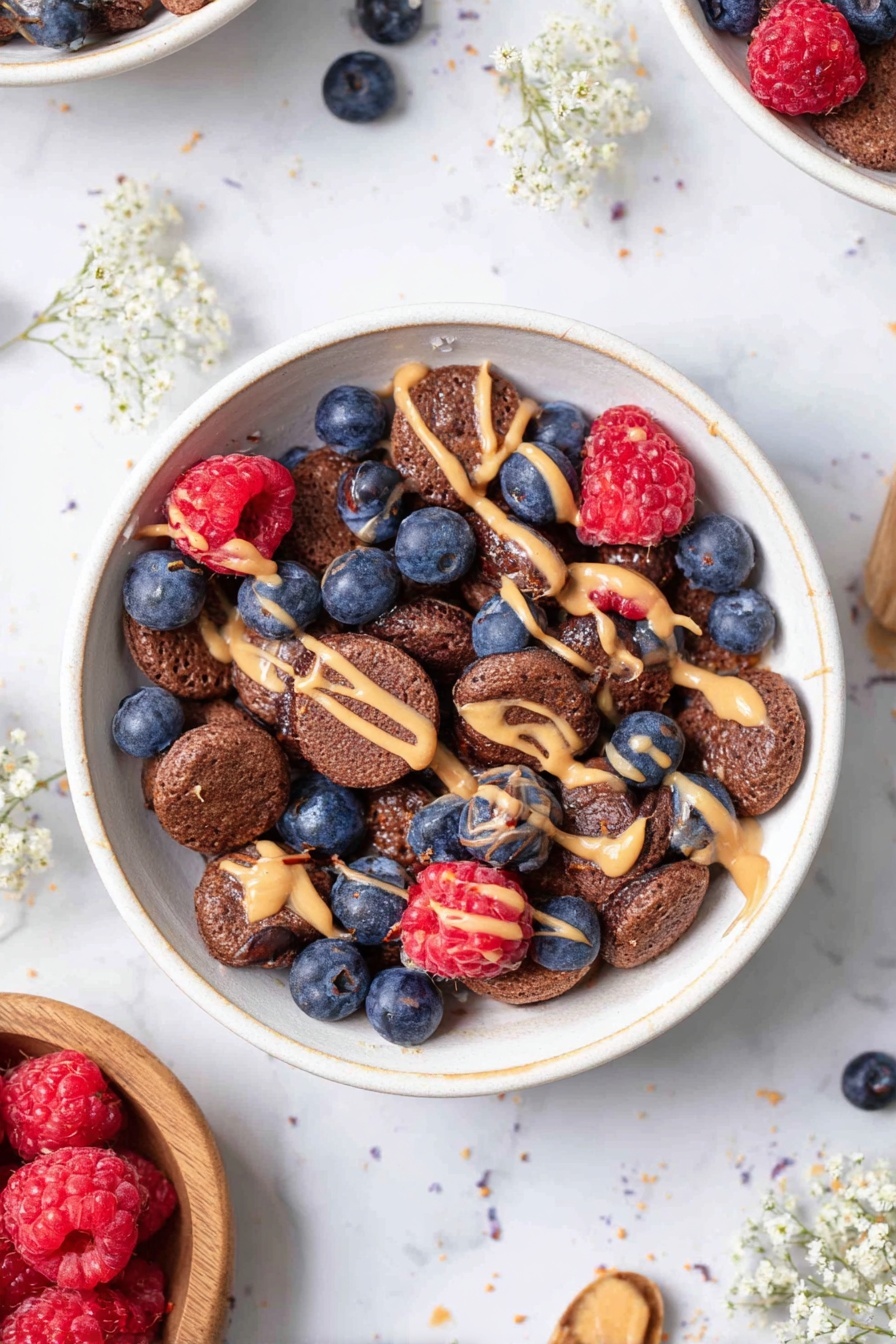 A white bowl filled with multiple small, round, dark brown pancake-like pieces forming the base layer; scattered on top are fresh blueberries and whole raspberries adding blue and red pops of color; thin streams of light brown peanut butter drizzle over the pancake pieces and berries, adding texture; the bowl is placed on a white marbled surface with some scattered blueberries and raspberries around, along with small sprigs of delicate white flowers; another bowl with similar contents can be partially seen at the top left and a wooden bowl with raspberries is at the bottom left; the scene is bright and clean, photo taken with an iphone --ar 2:3 --v 7 - Mini Pancake Cereal Bowl with Berries, pancake cereal recipes, adorable breakfast ideas, healthy pancake breakfast, berry breakfast bowls