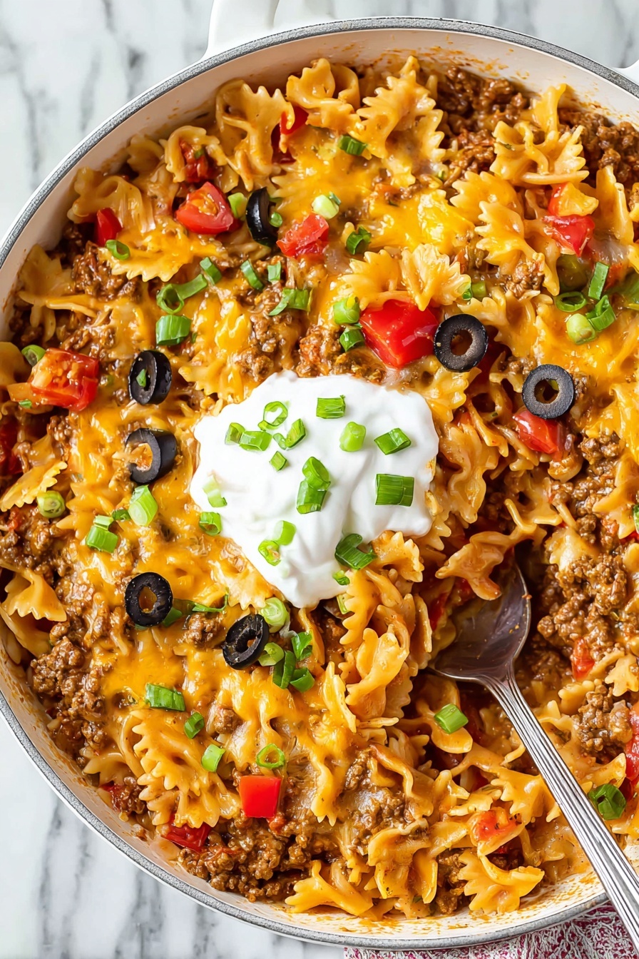 This dish is in a white frying pan filled with three main visible layers: the bottom layer is a mix of cooked ground meat and diced red peppers giving a brown and red base; on top of that, wavy-edged pasta pieces with a yellow-orange cheesy sauce are spread evenly; scattered on the surface are small green onion slices, black olive rings, and diced red tomatoes adding dots of green, black, and red. In the center, there is a dollop of white sour cream topped with a few green onion pieces. A silver spoon with pasta on it is seen inside the pan against a white marbled surface. photo taken with an iphone --ar 2:3 --v 7 - Cheesy Taco Pasta, easy taco pasta dinner, one-pot cheesy pasta, quick taco pasta recipe, flavorful taco pasta dinner